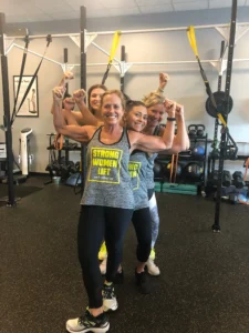Four women flexing their arms in a gym, wearing matching workout tops.